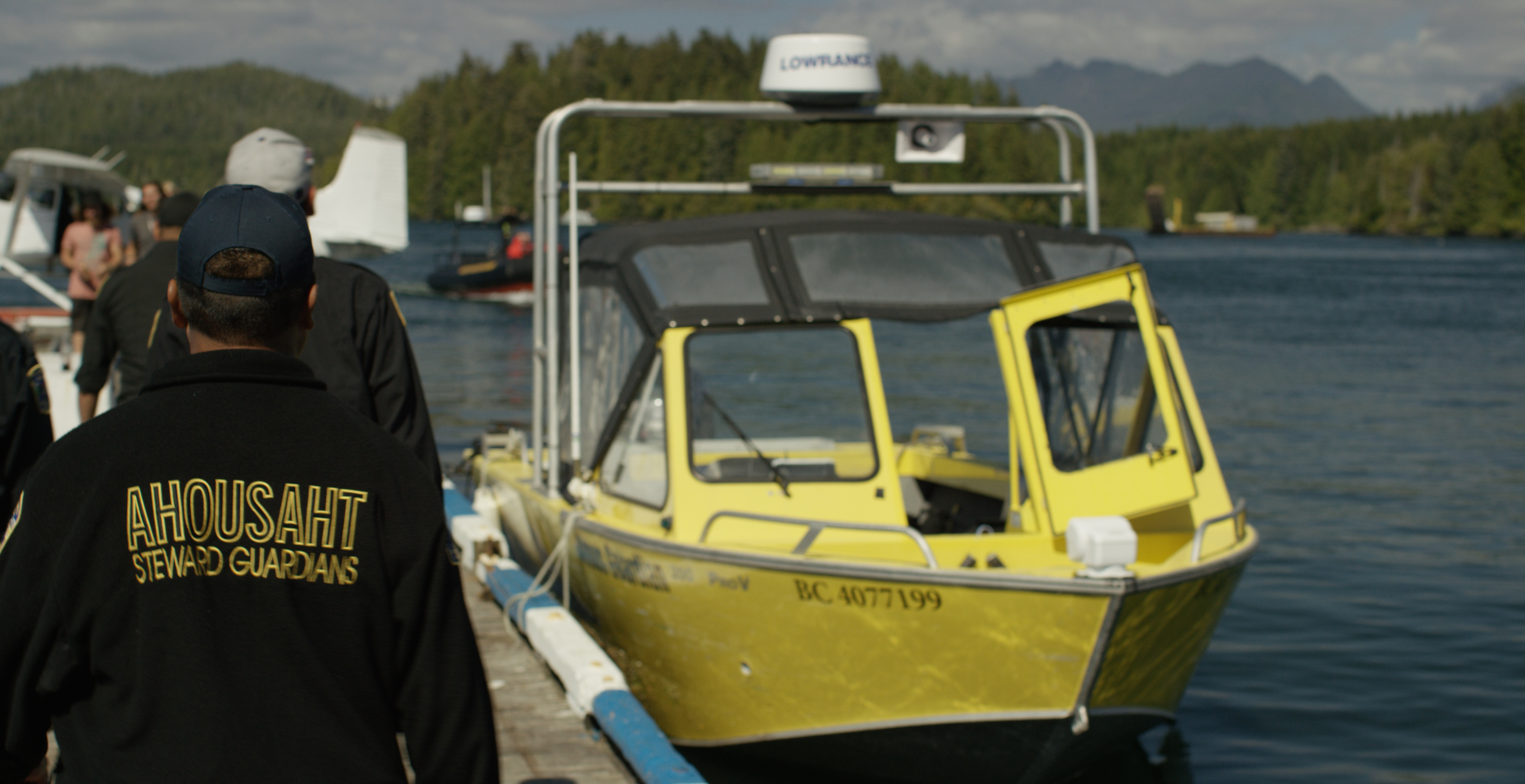 a man in a jacket that says, "ahousant stewardship guardians" walks away from the camera toward a boat