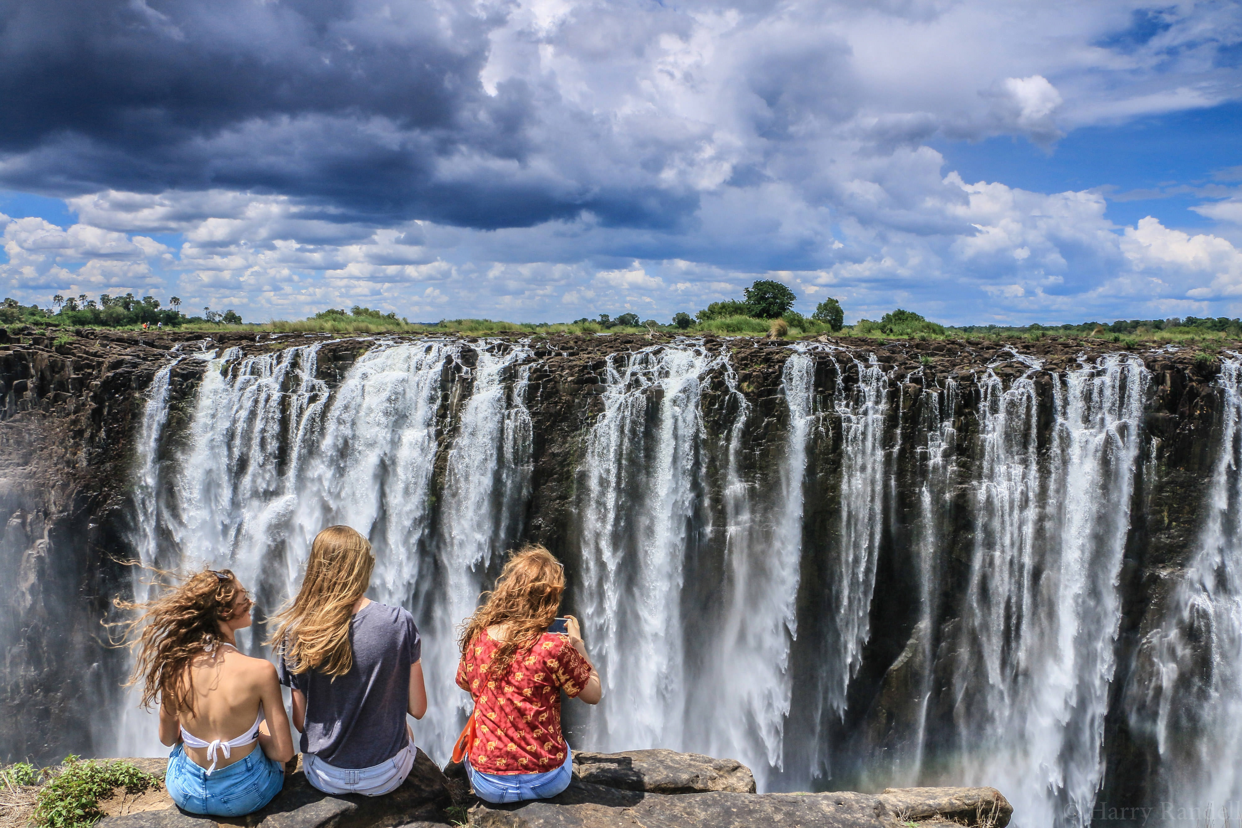 Three young people face Victoria Falls while their hair blows in the wind.